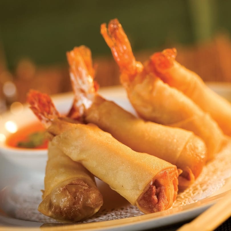 Crispy shrimp spring rolls with tails visible, served on a white plate with dipping sauce.
