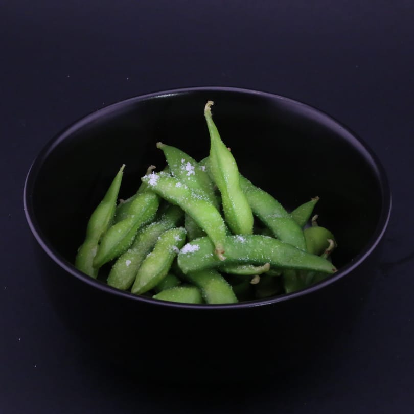 A black bowl filled with bright green edamame pods, lightly sprinkled with sea salt.