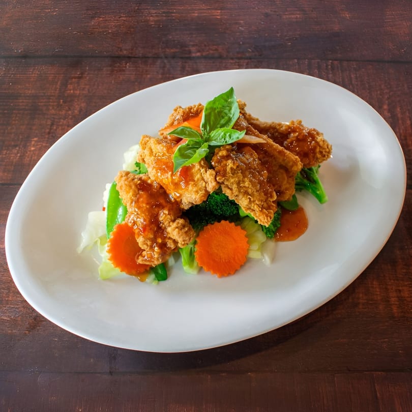 Crispy Thai chicken with sweet chili sauce, broccoli, carrots, and basil on a white plate.